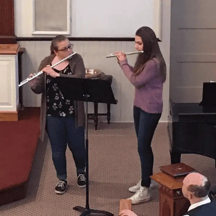 A close-up photo of two teenage girls playing flutes with a black music stand at the front of the church sanctuary.
