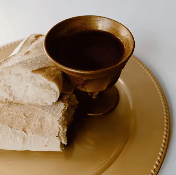 A carved wooden chalice of wine and a loaf of bread sliced in two stacked pieces sit on a brass-colored oval platter.