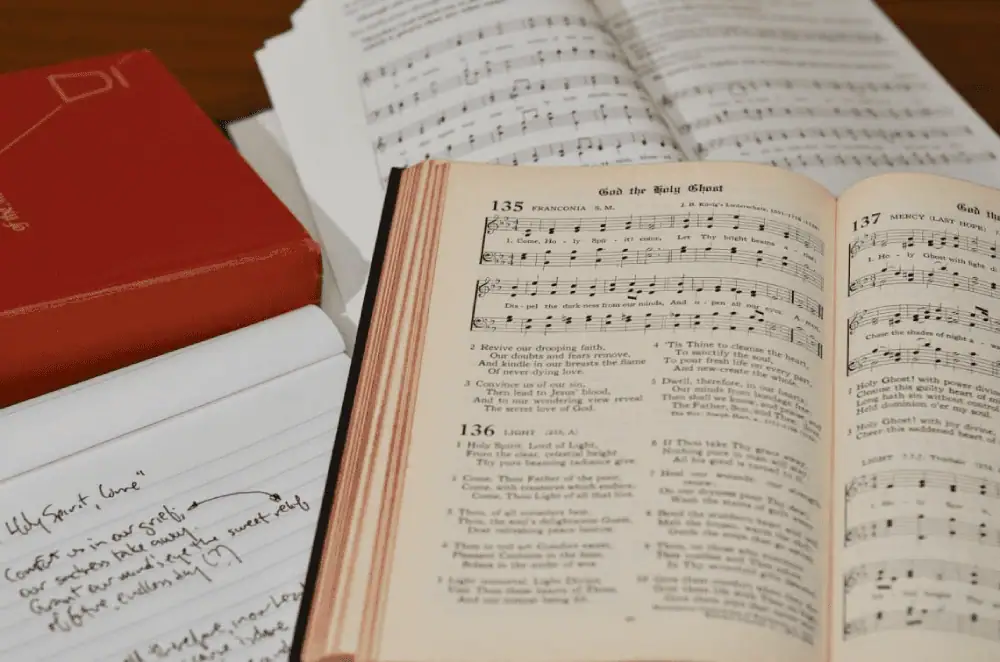 Three hymnals and a pad of lined paper are sitting on a table. A red hymnal is closed. Two hymnals are open displaying hymns, one of which has a black cover.