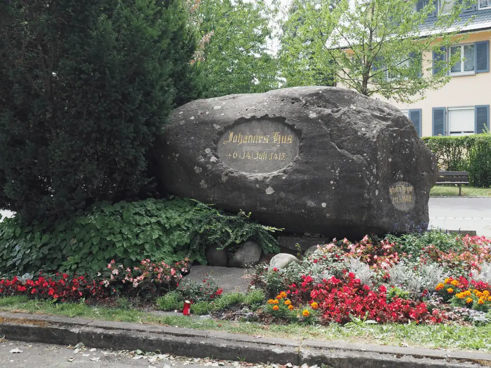 A large oblong stone made of black limestone marks the place where Jan Hus is believed to have been martyred. The stone has an inscription in gold lettering, reading "Johannes Hus + 6 Juli 1415". There is a large evergreen tree to the left of the stone, and it is surrounded by spring flowers in colors of red, pink, yellow, white, periwinkle, and orange.