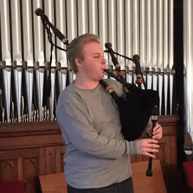 An older teenage boy plays a set of black bagpipes in front of the organ chamber in the balcony at Schoeneck.