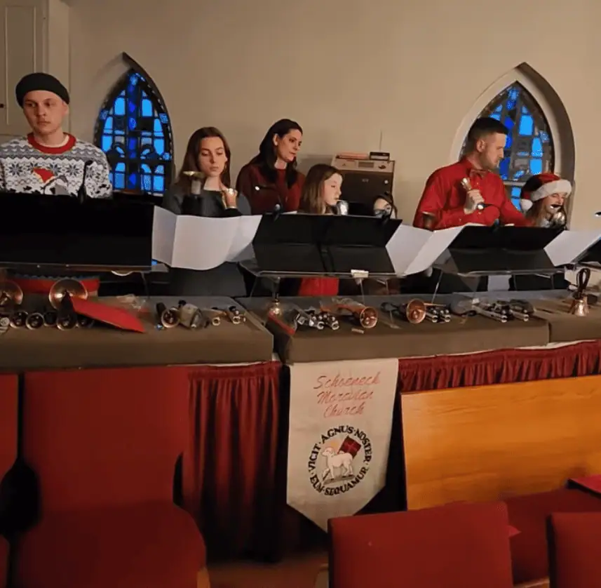 A photo of the Junior Bell Choir as they play in the balcony on Christmas Eve. There are five young people and kids playing the bells with two adult helpers. One of the youth is wearing a Santa hat. The bell tables have grey cushions on top with burgundy table skirts. There is a banner in the middle with Schoeneck Moravian Church and a Latin Moravian Seal cross-stitched on it.
