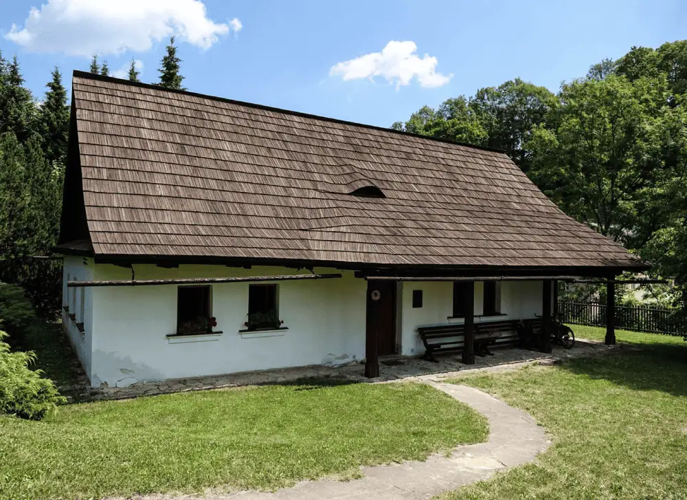 A photo of the "Na Sboru" house in Kunvald where the Unity of the Brethren gathered in the 1450s. It is a small, rectangular, white building, with a steep brown shingled roof, surrounded by grass and trees on a sunny day.