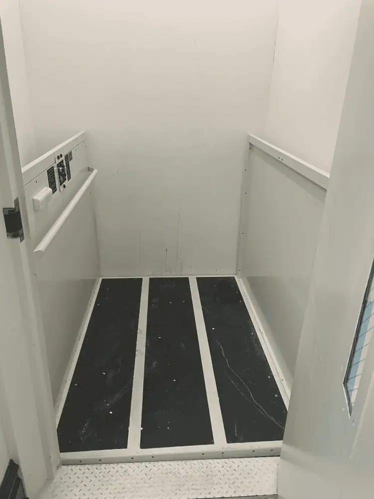 A photo of the inside of the lift. It is white with a handrail. The floor has three wide stripes of black anti-slip material.