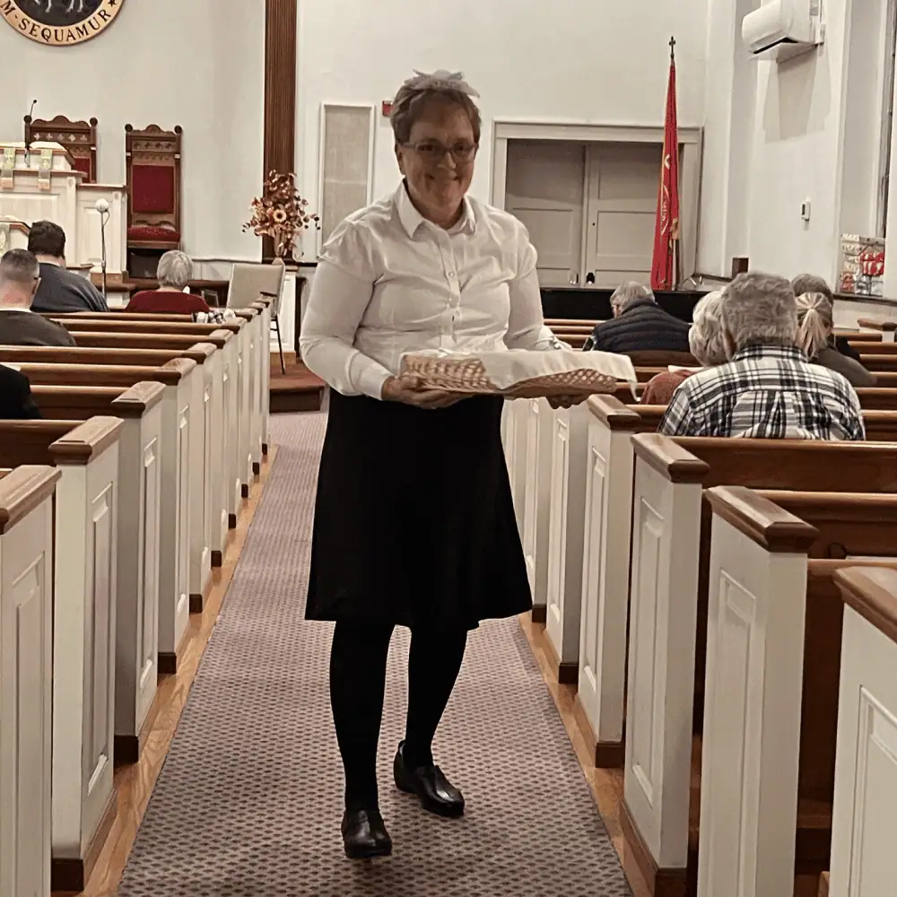 A woman dressed in a white blouse with a black skirt, stands in the church aisle holding a basket of sugar cake.