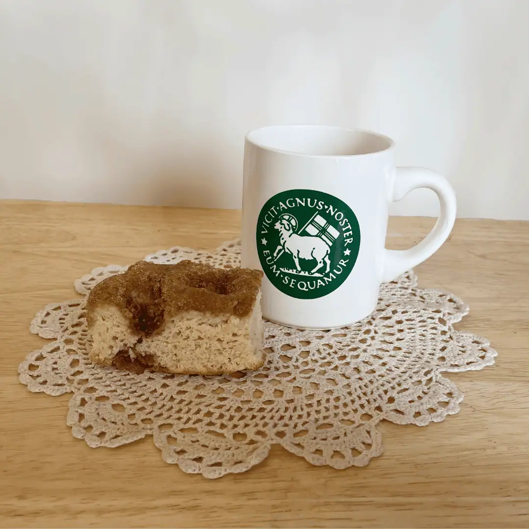 A white coffee mug with a green Moravian seal and a piece of sugar cake sit on a crocheted doily on a light pine tabletop.
