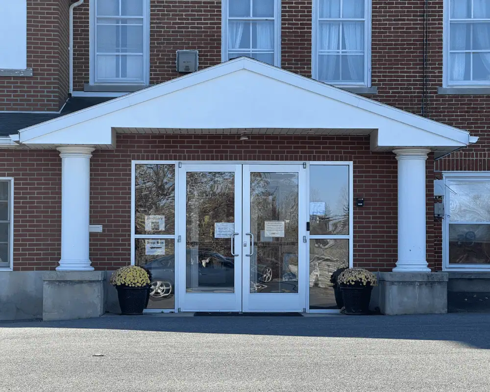 A photo of the main entrance of Schoeneck Moravian Church from the parking lot. The building is made of red brick and the entrance is covered with two white posts at either end. Next to the posts are black planters with yellow flowers. There is a glass double door with full length windows on each side.