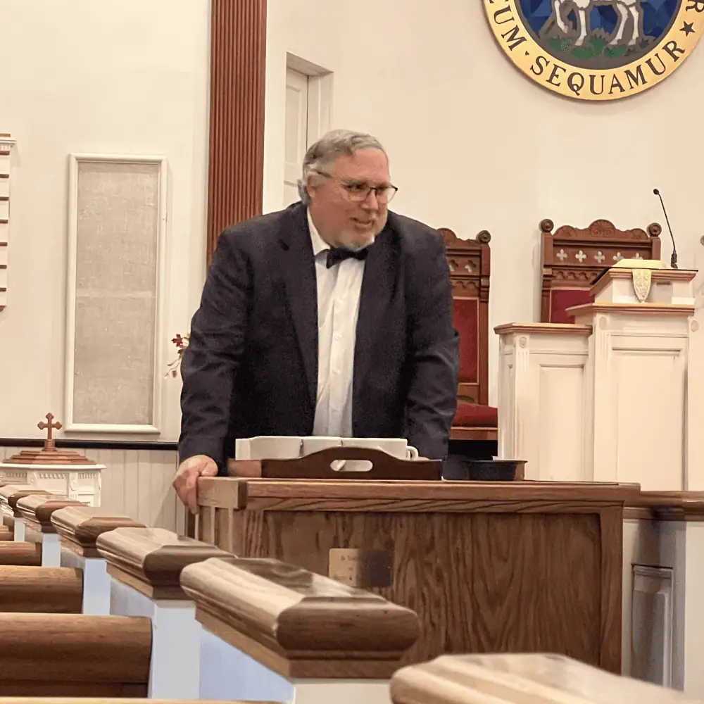 One of our Diener men stands in the sanctuary with a lovefeast cart. He is wearing a black suit with a white dress shirt and a black bowtie. On top of the cart is a tray of coffee mugs.