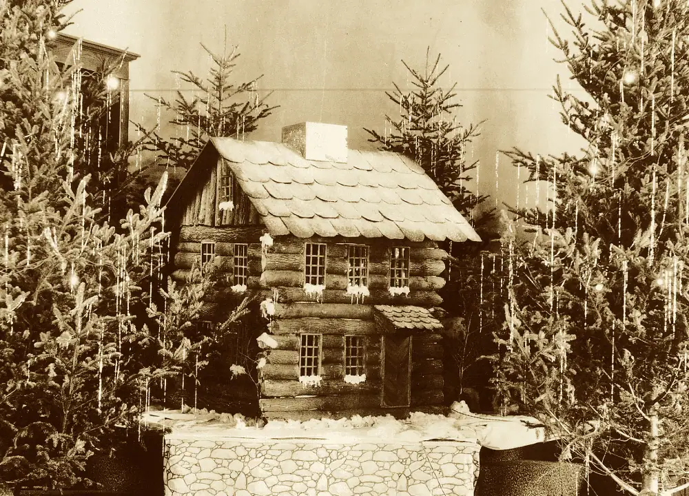 A sepia-toned photograph of a model of the original log church. The model has two stories, the first floor has two windows on the right and a covered door on the left. The second floor has three windows across the building. The roof looks to be in the style of round wooden shingles. There is snow on each of the windowsills. The model is surrounded by small live Christmas trees with twinkle lights and strands of silver tinsel.