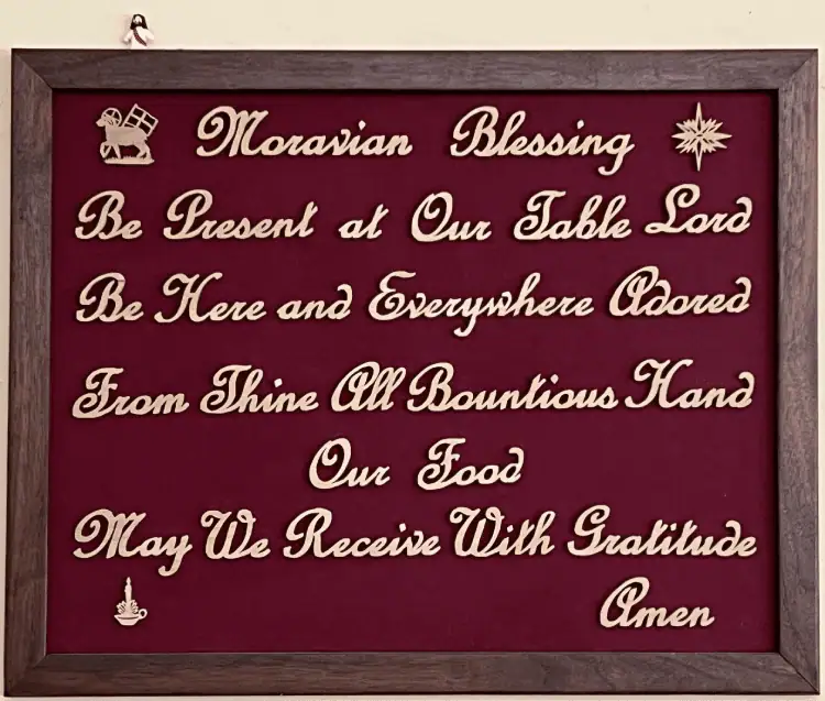 A Moravian Blessing that hangs in the Fellowship Hall at Schoeneck. The frame is made from a dark wood and it has a burgundy fabric background. The words of the blessing are carved from a thin light-clored wood. There is also an Agnus Dei, a Moravian star, and a lovefeast candle carved from the same light-colored wood. The Moravian Blessing is: "Be present at our table Lord, Be here and everywhere adored, From thine all bounteous hand our food, may we receive with gratitude." (There is also a very tiny plastic figure of Jesus standing on top of the frame.)
