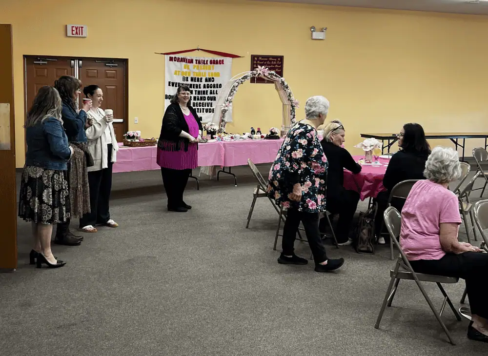 There are a number of women in the Fellowship Hall, which is decorated for the annual Mother's Day tea. There are two folding tables for a buffet, with pink tablecloths and a decorative floral arch. The round tables also have pink tablecloths. Some of the women are standing, ready to go through the buffet and some of the women are seated at the tables.
