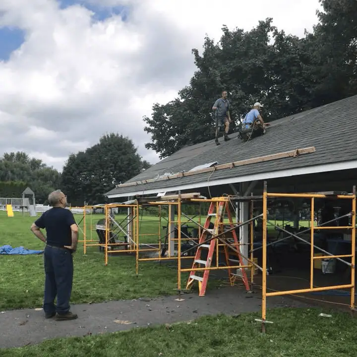 A photo showing the church pavilion with the roof being replaced. The sky is blue with big white puffy clouds. A man stands on the ground looking up next to bright yellow scaffolding that spans the length of the pavilion. There are two men working on the roof, one kneeling and one standing.