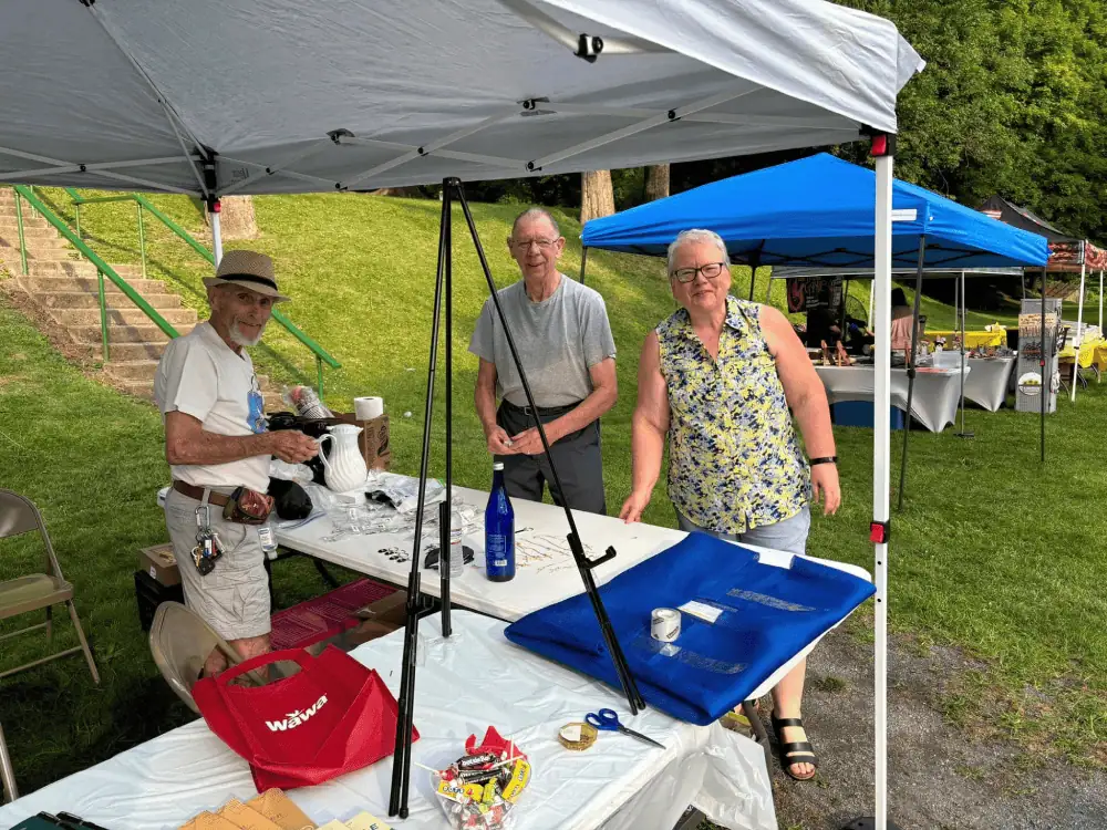 Three Schoeneckians stand under a white tent while setting up their display for the annual Nazareth Day celebration in the park.