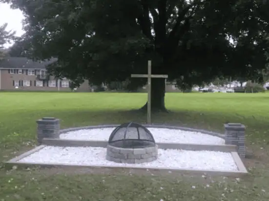 A close-up photo of the current fire pit at Schoeneck. There are two connected areas - a rectangular area filled with white stone which has a circular firepit made of large grey bricks with a steel screen dome cover in the middle. The other area is curved, also filled with white stone, with a wooden cross standing at the end. Two grey stone pillars stand at the side edges where the two sections meet.