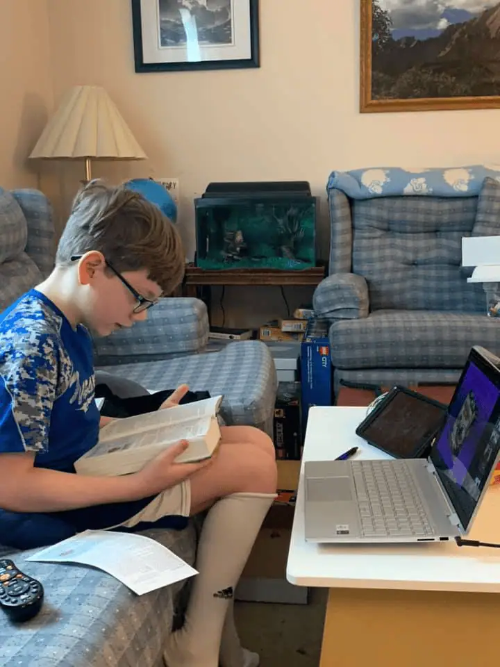 A photo of a boy sitting on the couch in his living room. The couch is made of fabric with checks in shades of light blue and darker blue. On a white table in front of the boy is an open laptop. The boy is holding an open Bible on his lap and he is looking down at it. Next to him is a white paper with a lesson printed on it.