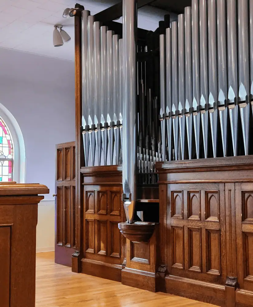 A view of the organ pipes in the balcony. The hardwood floor is a medium colored wood. The organ cabinet is a dark colored wood and the organ pipes are a gleaming silver.