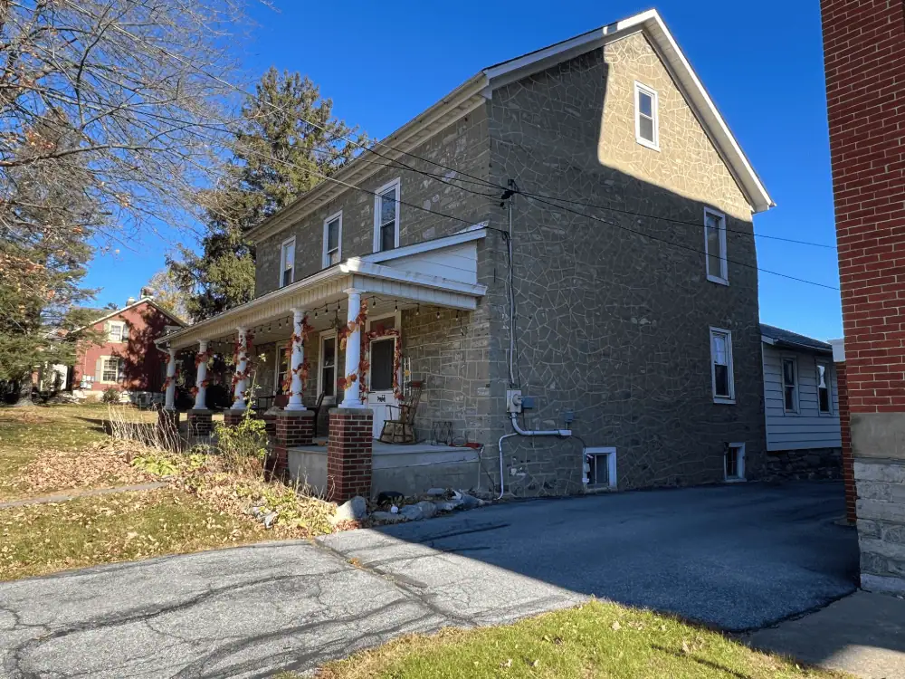 A picture of the front and right of the parsonage on a sunny day. The parsonage is a light colored stone building with white trim around the windows and front door. The porch is cement with five pillars that are half brick and half round white posts. The posts are decorated with strands of fall colored faux leaves wrapped around them The front lawn is strewn with fallen leaves. Along the side of the parsonage, you can see a white sided addition.