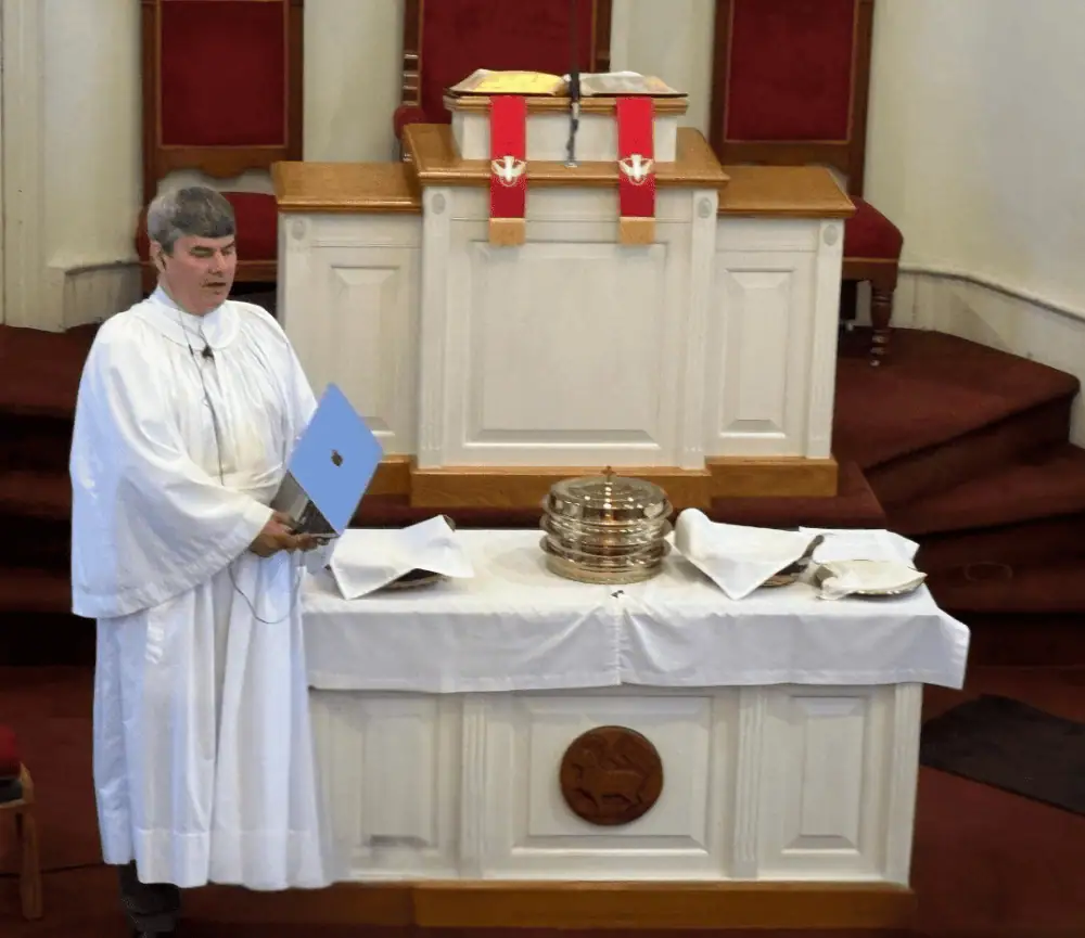 Pastor Ian wears his white surplice as he stands next to the communion table. The communion table has a white tablecloth with the communion elements on top.