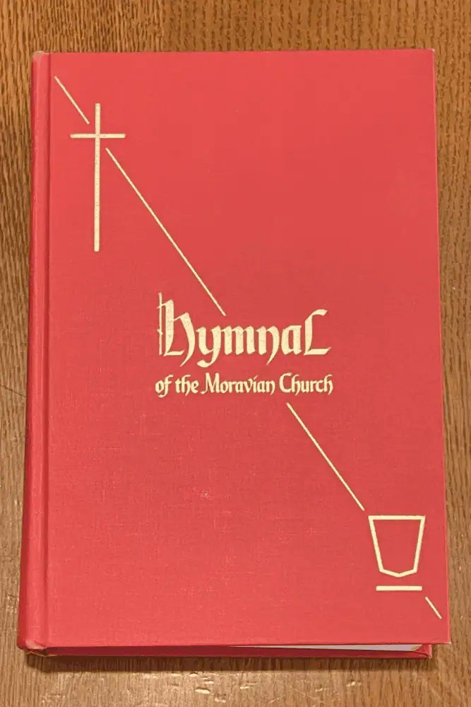 A photo of the cover of the red hymnal, entitled simply as "Hymnal of the Moravian Church" in gold embossed lettering. At the top left corner is a gold embossed cross, with an embossed line leading diagonally to the title. A gold embossed line then leads to the lower right corner and a gold embossed image of a simple communion chalice.