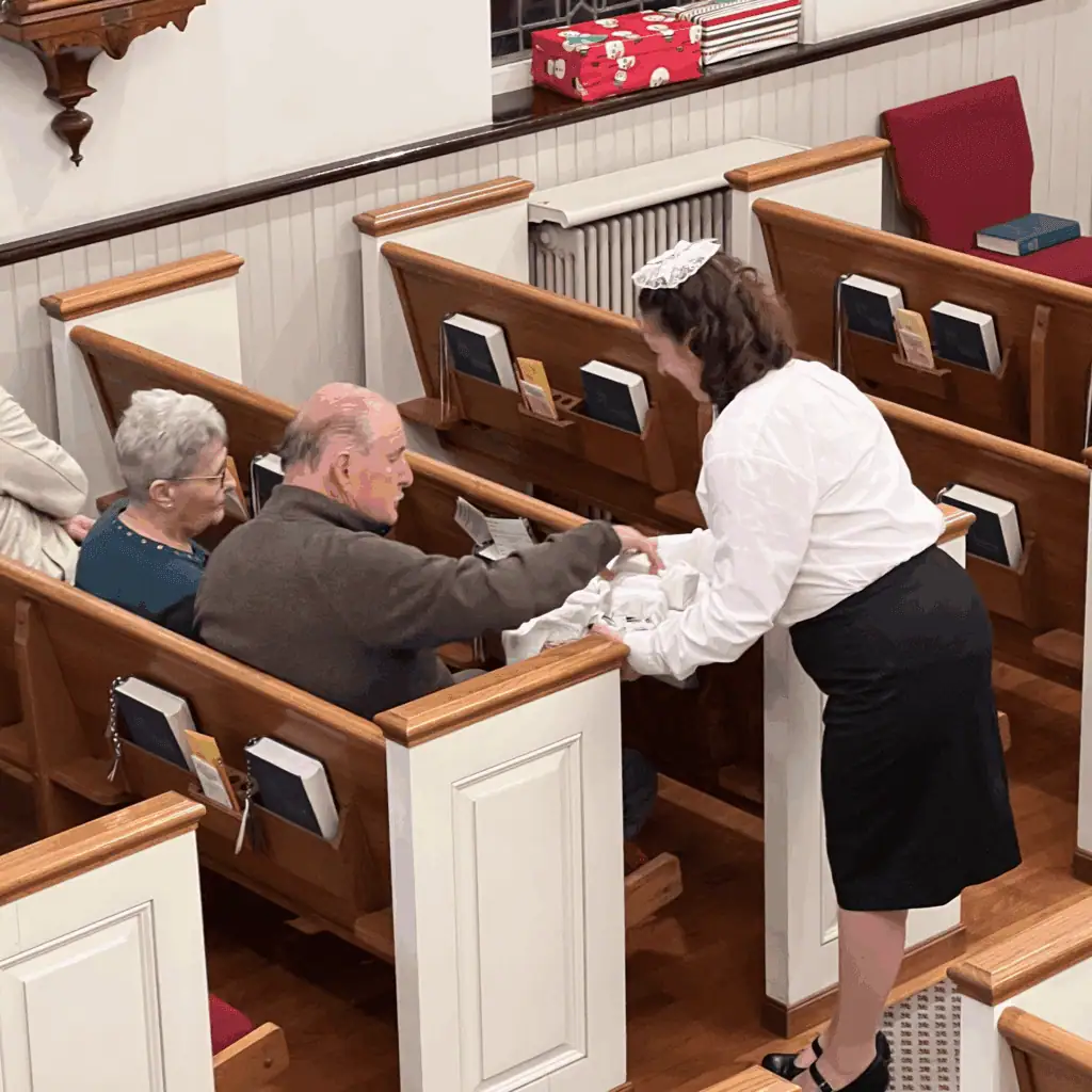 A woman dressed in a white blouse with a black skirt, wearing a traditional lace hauba in her hair, serves sugarcake to two congregation members as they sit in the pew.