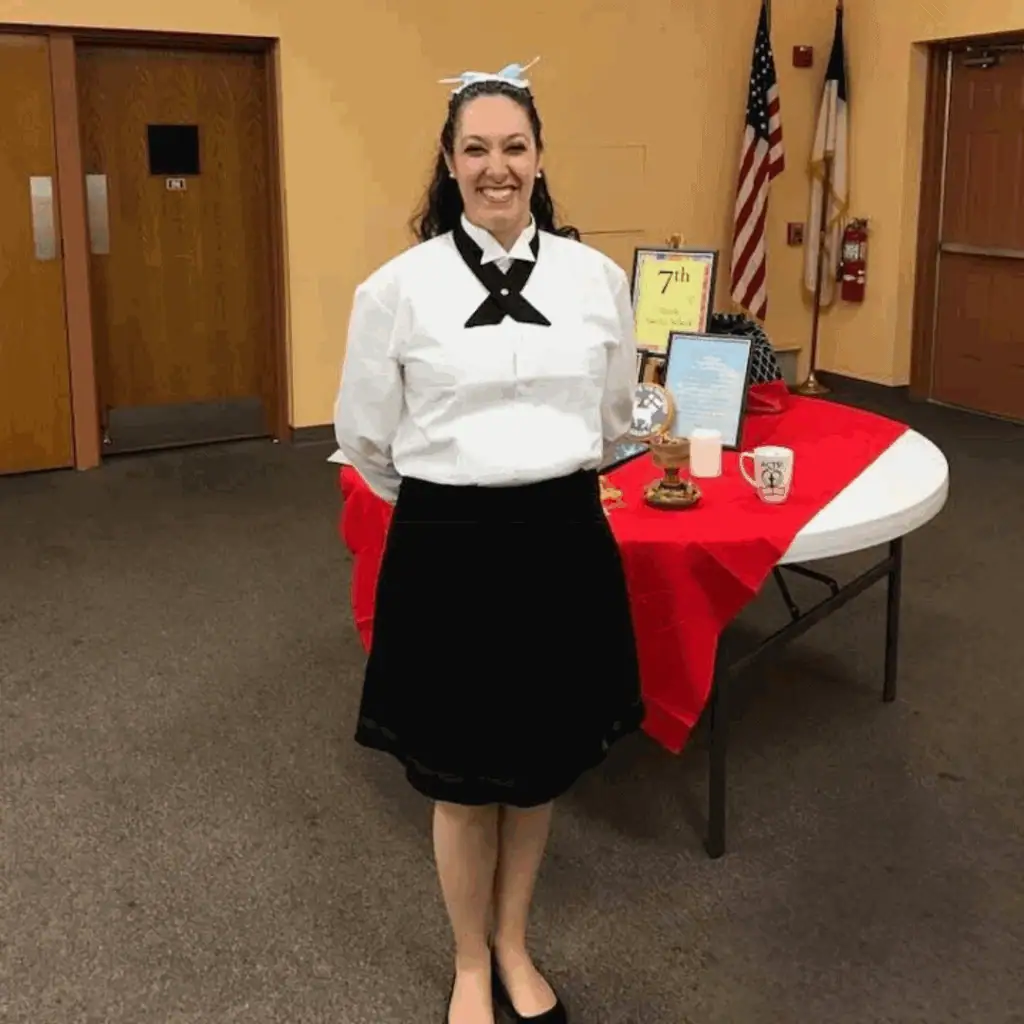 A smiling young woman stands in the Fellowship Hall, ready to serve with the Diener Corps. She is wearing a black skirt with a white blouse and a black cross-over tie. On her head is a traditional hauba with light blue ribbon, indicating she is a married woman.