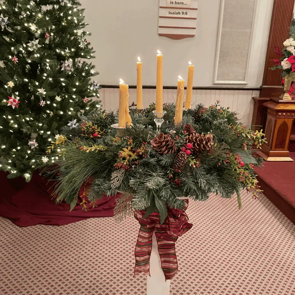A photo of Schoeneck's Advent Wreath. In the background on the left is the sanctuary Christmas tree decorated with white lights and multi-colored paper Moravian stars. The wreath has 5 beeswax candles, lit for Christmas Eve. The candles are surrounded by a few different types of evergreen branches, with pine cones and red berries mixed about. The wreath sits on top of a white stand, which has a fabric bow - with red, green, and sheer white stripes - tied around it.