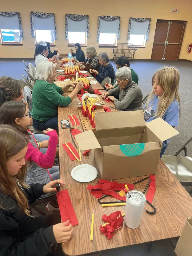 An intergenerational group of people sit at two brown folding tables wrapping red frills around beeswax candles in preparation for Christmas Eve services.