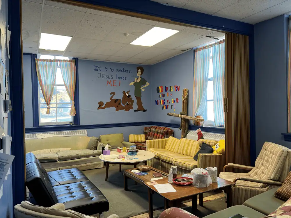 The Senior Youth Room has several comfortable couches, chairs, and a futon lining the walls. There are three coffee tables spaced among the furniture. The walls of the room are light blue and the two windows are trimmed in navy. On the wall is a mural of Scooby Doo and Shaggy with the words "It's no mystery - Jesus loves me!"
