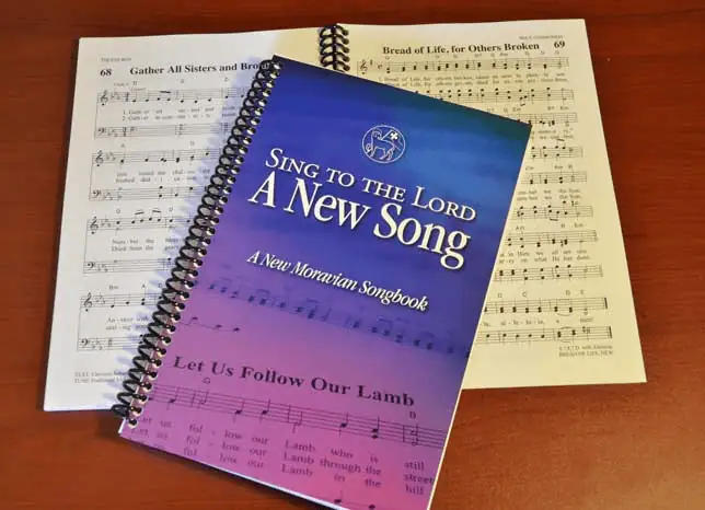A closed hymnal "Sing to the Lord a New Song - A New Moravian Songbook" lies on top of an open hymnal of the same. The cover of the closed hymnal has a black spiral binding and is a dark blue at the top and fades through to a bright purple at the bottom.