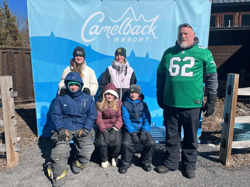 Six Schoeneckians - five youth and one brave adult - are bundled up for winter weather in front of a light blue banner that reads "Camelback Resort"