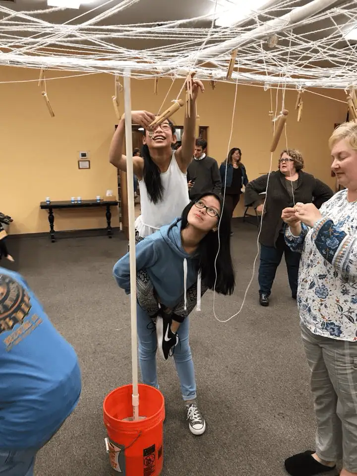 During the annual Spider Web Social, two sisters work together to untangle the string "web". The older sister is holding her younger sister on her back piggy-back style while a woman looks on.