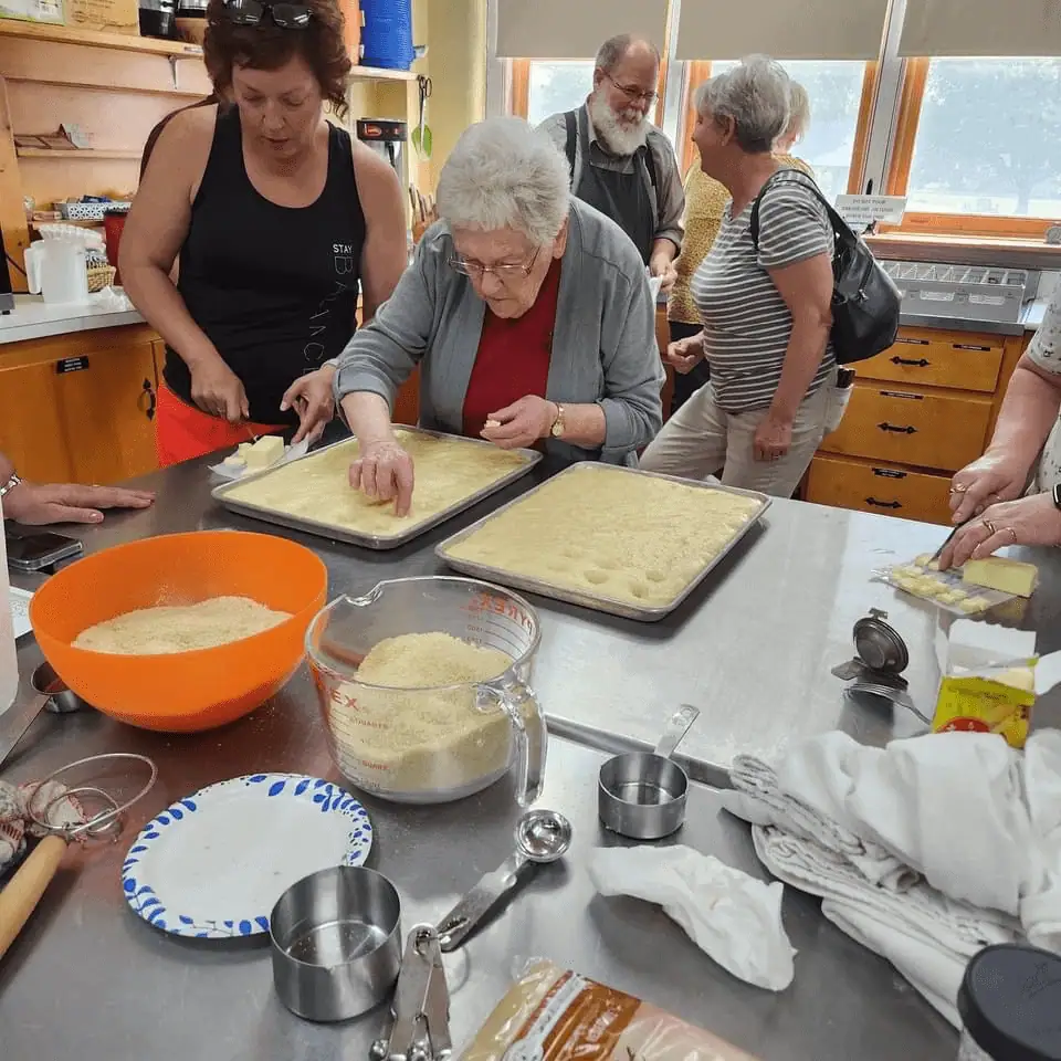 In the kitchen at Schoeneck, several women and men are baking sugarcake for the congregation. On the large stainless steel work surface are measuring cups, mixing bowls, and two baking trays of sugarcake dough. Two women are working together - one pokes holes in the dough with her fingers while the other fills the holes with a butter and brown sugar mix.