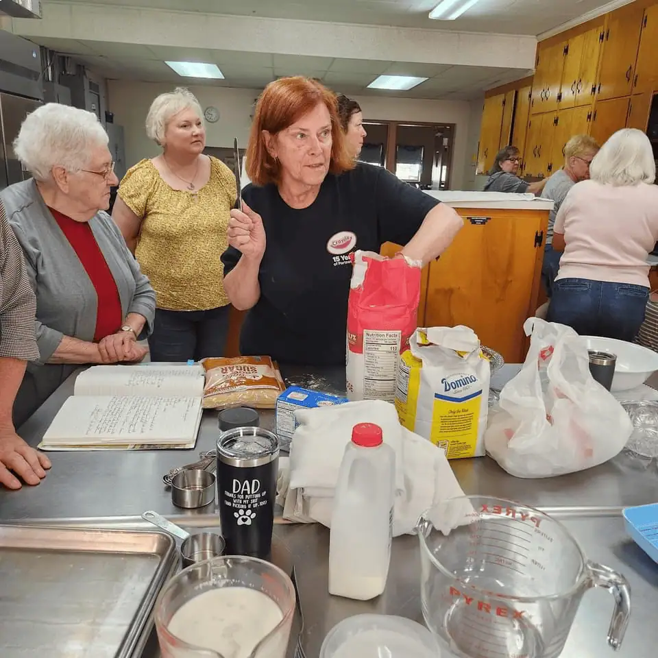 Seven Schoeneckians stand around the prep and cook area of Schoeneck's kitchen. On the stainless steel island is a cookbook, and ingredients for making sugar cake, along with various cooking implements.