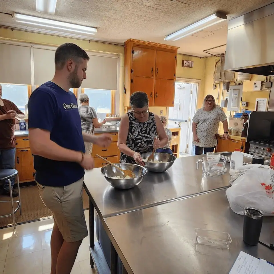 Five Schoeneckians stand in the prep and cook area of the Schoeneck kitchen. A man and a woman are next to the stainless steel island, stirring ingredients in large stainless steel bowls.