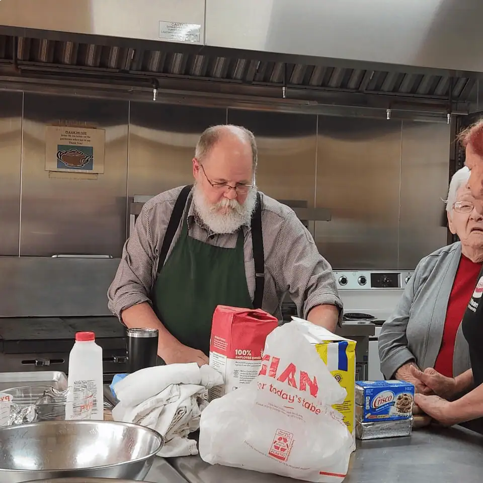 A man with a beard stands at the stainless steel island in the prep and cook section of Schoeneck's kitchen with two women, showing them the ingredients on the island.