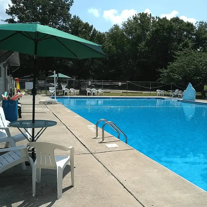 A photo of the pool at Camp Hope. The water in the empty pool is an invitingly aqua blue color. Along the concrete pool deck in the left corner of the photo is a small table with a green umbrella and three white plastic chairs,
