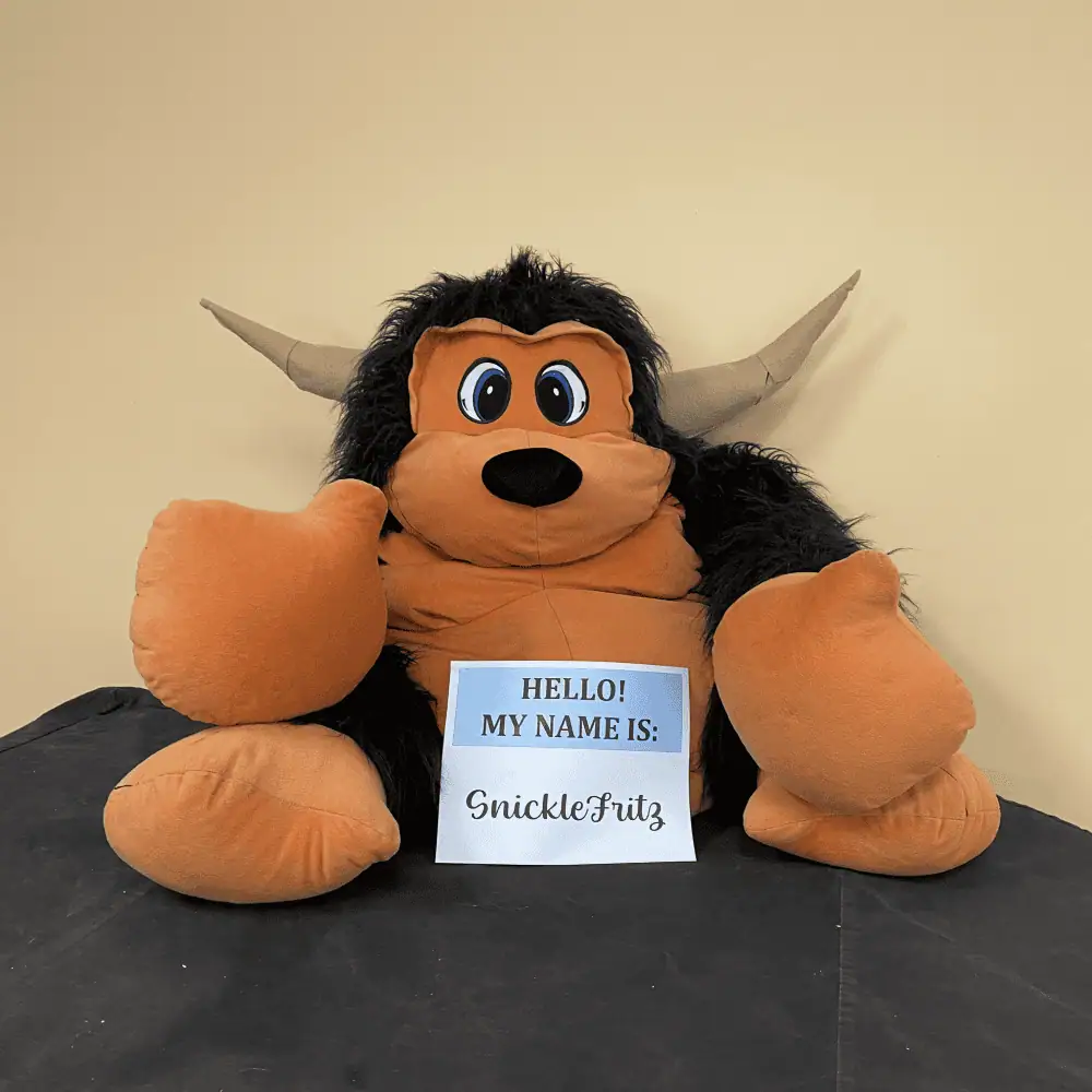 A picture of the stuffed yak who is the mascot for our "Snack 'N Yak" fellowships. He is a burnt umber color with black fur on his arms, legs, and head. Two grey horns point out sideways from his head. A piece of paper in front of him introduces him as Snickle Fritz.