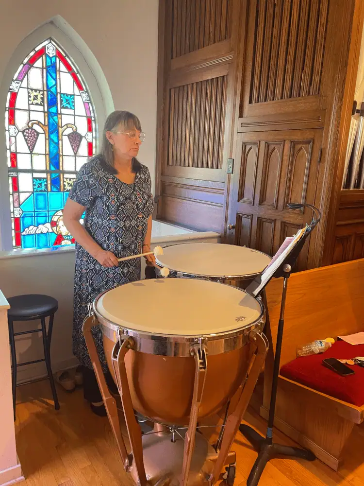 An older women is playing two timpani drums in the church balcony. In front of the copper-colored drums is a black music stand which is next to a wooden pew with a red seat cushion. Behind the woman is one of the stained glass windows of the church, and to her right is the door for the pipe organ chamber.