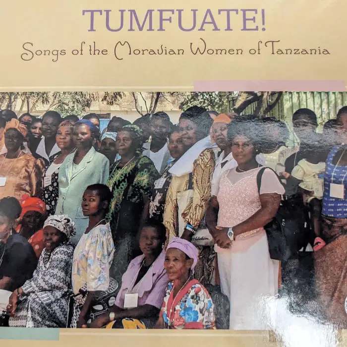 The title of the CD is at the top with "Tumfuate!" in large all caps lavender serif letters. The subtitle "Songs of the Moravian Women of Tanzania" is written in black underneath in a modern sans serif rounded font. Beneath the title is a large group of Tanzanian women of different ages in a variety of styles of dress. Some women are wearing dresses with traditional African patterns, and some are wearing more contemporary style dresses. Some of the women are wearing nametags around their necks.