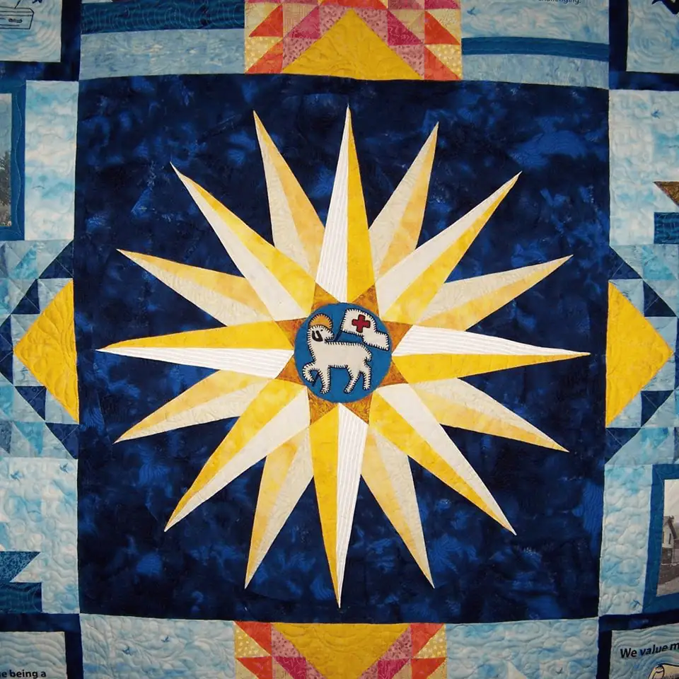A close-up photo of a quilt square showing a Moravian star. The star's points are made of white and yellow fabric with a stitched Agnus Dei in the middle of the star. The star is on a field of dark blue fabric with slightly lighter blue splotches. The quilt square is framed by strips blue fabric with white areas that look like clouds.