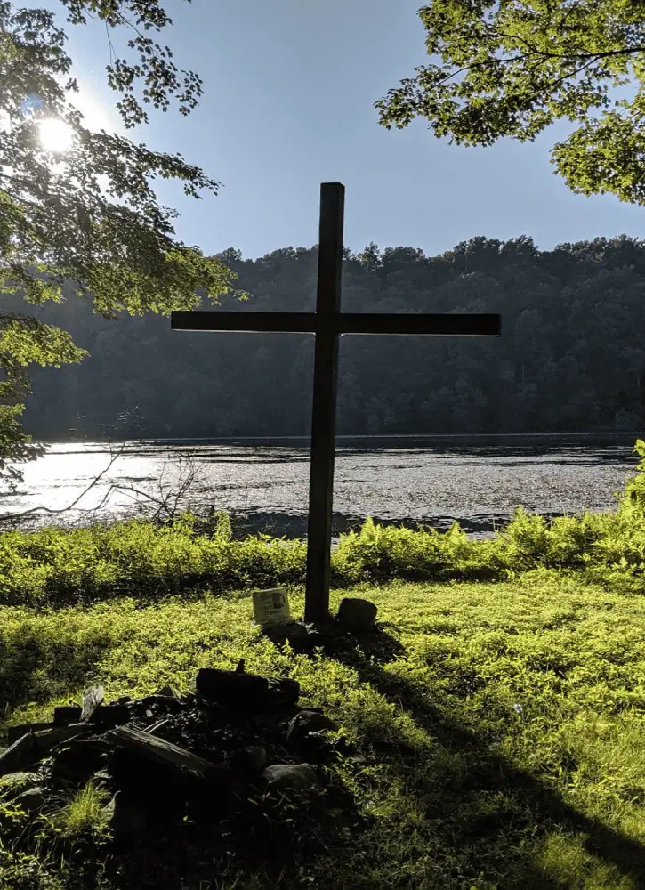 A photo of a rustic cross stands at the edge of Little Silver Lake. The sun is behind the cross above the blue-grey water. The grass is green and the cross casts a long shadow across it. In the foreground is an empty campfire circle.