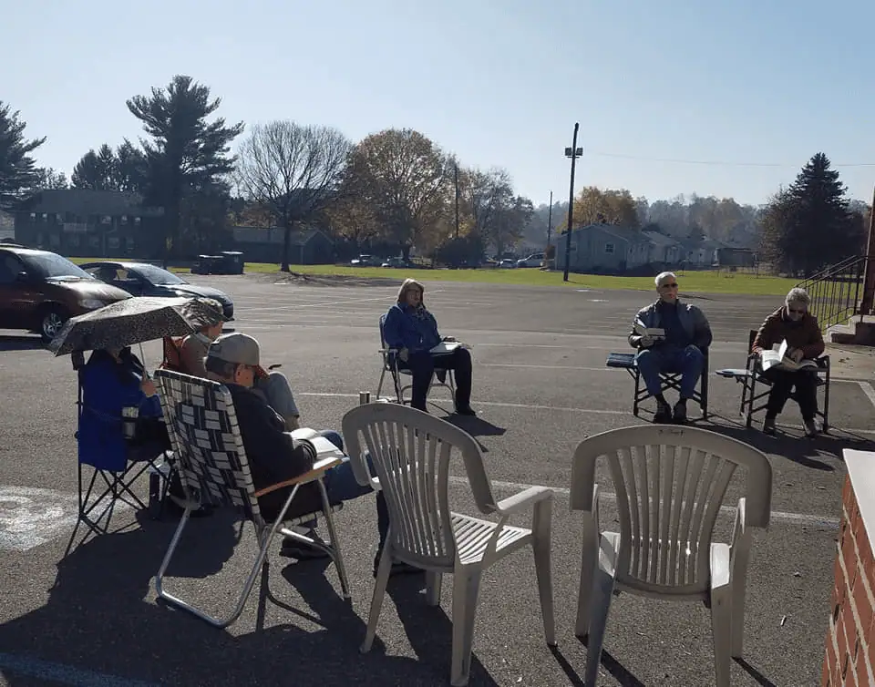 A picture of several adults sitting in lawn chairs in Schoeneck's parking lot. The chairs are separated in a few groups. One person is holding an umbrella for shade from the sun.