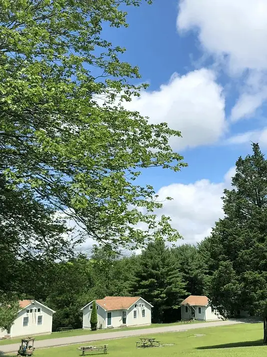 A photo of Camp Hope shows three year-round cabins from a distance. The cabins are a beige color with light brown roofs. The sky is blue with puffy white clouds. Tall trees are behind the cabins and a field with a few picnic tables are in front of the cabins.
