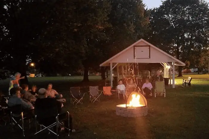 A color photo of a Fire Pit Fellowship evening. A number of people are sitting in lawn chairs around a fire pit made of grey stone bricks. There is a bright fire burning, with steel mesh half-circle lid on top of the fire pit. The pavilion is just behind the people sitting around the fire.