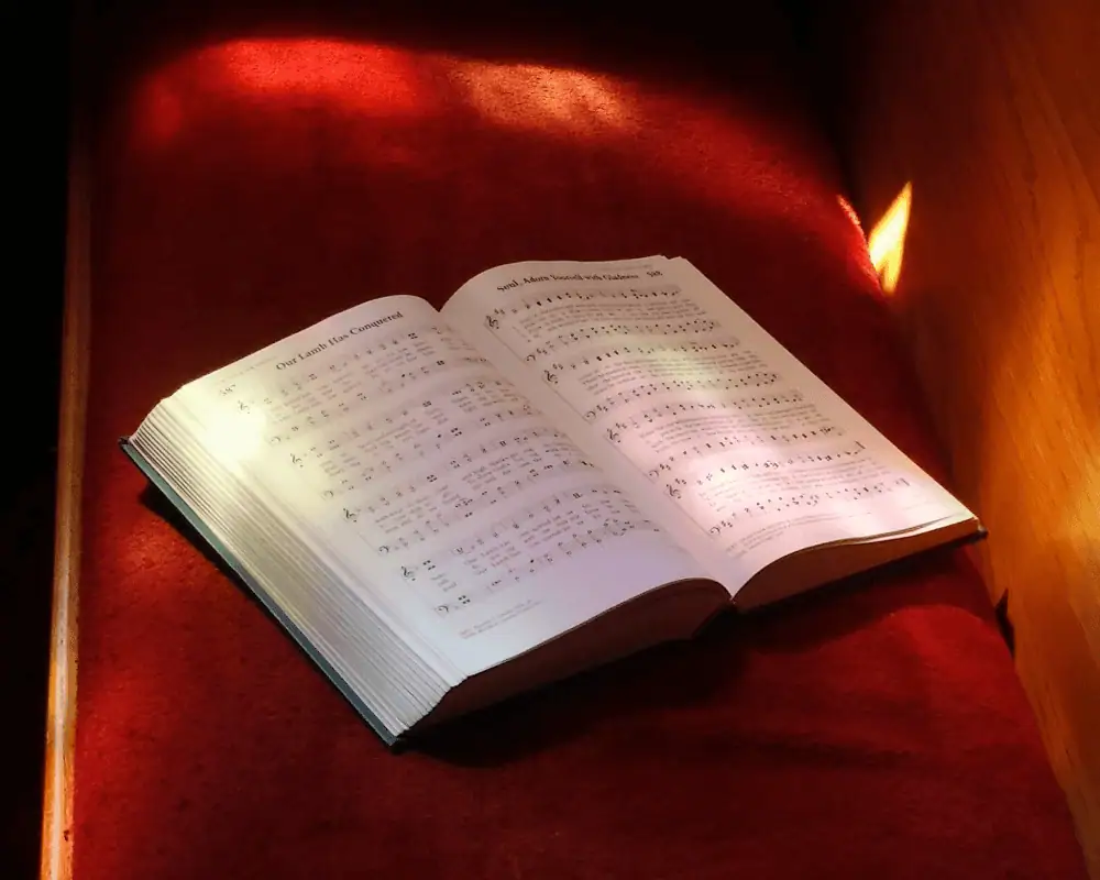 A close-up photo of an open hymnal resting on one of the pews in the sanctuary. The late afternoon sun shines through a stained glass window and onto the hymnal and pew.