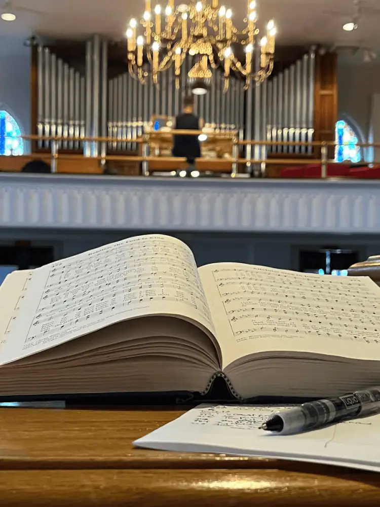 A photo taken from the vantage point of Schoeneck's pulpit looking towards the balcony at the back of the church. In the foreground is a close-up of an open hymnal on the book stand with a piece of paper with handwritten notes, and a black pen. The photo looks up towards the balcony, which is out of focus. The chandelier and most of the balcony with the organ pipes are visible, and a man is at the organ console.