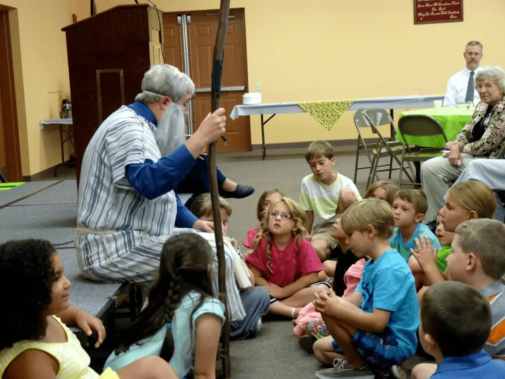 A photo taken during a summer worship service in the Fellowship Hall at Schoeneck. Pastor Terry sits on the edge of a low grey platform. He is wearing a thin white robe with grey stripes over his suit, and a long, fake grey beard while holding a wooden shepherd's crook. A number of children are crowded around him, sitting on the floor as he addresses them, while the congregation looks on.