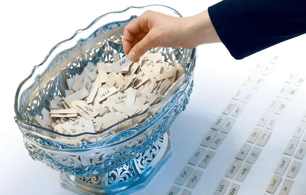 A large wide decorative silver and glass bowl on a white table holds small numbered scraps of paper. A hand reaches into the bowl to choose one of the pieces of paper, and there are three rows of chosen papers on the table in front of the bowl.