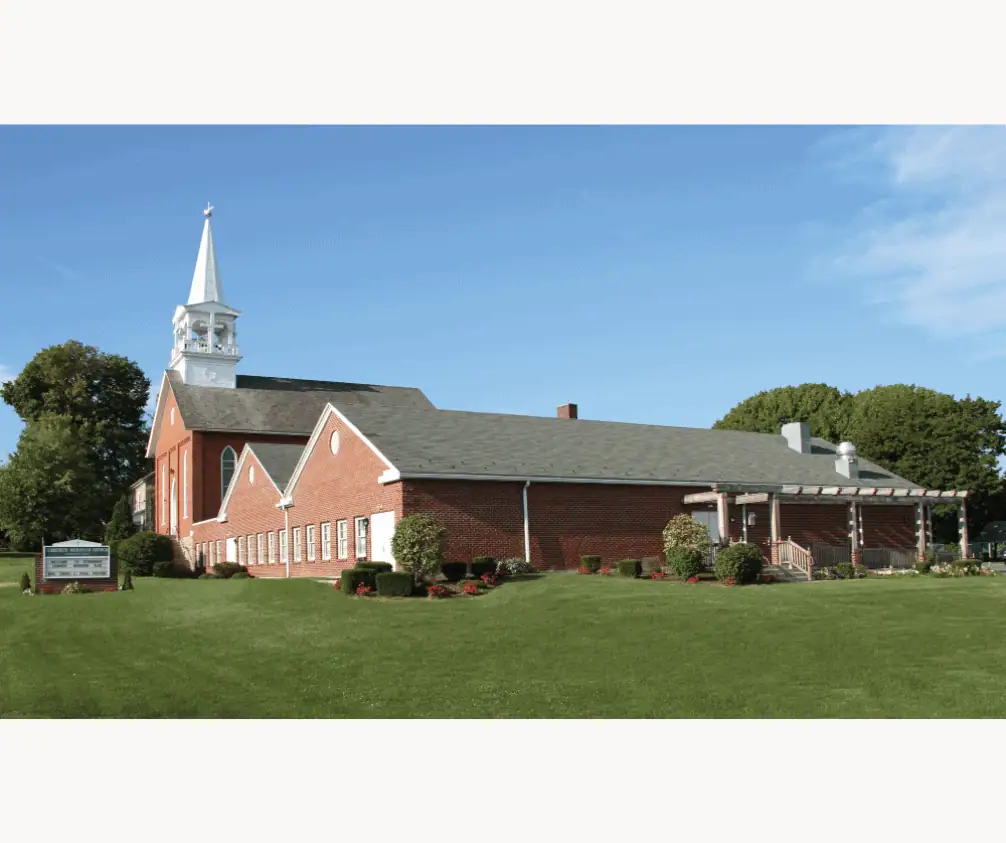 This photo shows a view of Schoeneck Moravian Church from a southwestern vantage. The red brick church sanctuary building is at the top of the photo, with the 1997 Fellowship Hall addition towards the center of the photo. The old church sign with hand-added lettering is visible to the left edge of the photo. Along the side of the building, the pergola outside the Fellowship Hall is visible. The picture was taken on a bright sunny day with blue skies and trees in full bloom. The green church lawn is in the foreground of the picture. This photo shows a view of Schoeneck Moravian Church from a southwestern vantage. The red brick church sanctuary building is at the top of the photo, with the 1997 Fellowship Hall addition towards the center of the photo. The old church sign with hand-added lettering is visible to the left edge of the photo. Along the side of the building, the pergola outside the Fellowship Hall is visible. The picture was taken on a bright sunny day with blue skies and trees in full bloom. The green church lawn is in the foreground of the picture.