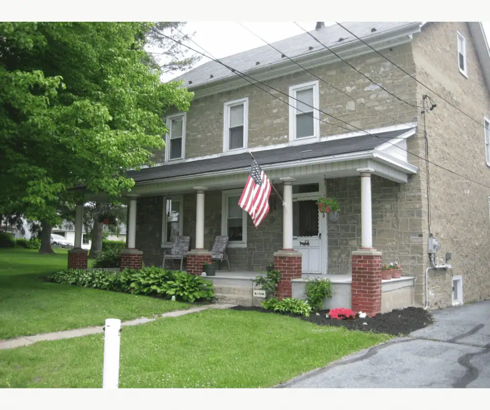 A picture of the front of the parsonage on a cloudy day. The parsonage is a light colored stone building with white trim around the windows and front door. The porch is cement with five pillars that are half brick and half round white posts. An American flag is hanging from one of the posts. The grass is green and the tree in front is in full bloom. The small garden along the front of the porch is filled with several hosta and a small pink azalea. A picture of the front of the parsonage on a cloudy day. The parsonage is a light colored stone building with white trim around the windows and front door. The porch is cement with five pillars that are half brick and half round white posts. An American flag is hanging from one of the posts. The grass is green and the tree in front is in full bloom. The small garden along the front of the porch is filled with several hosta and a small pink azalea.
